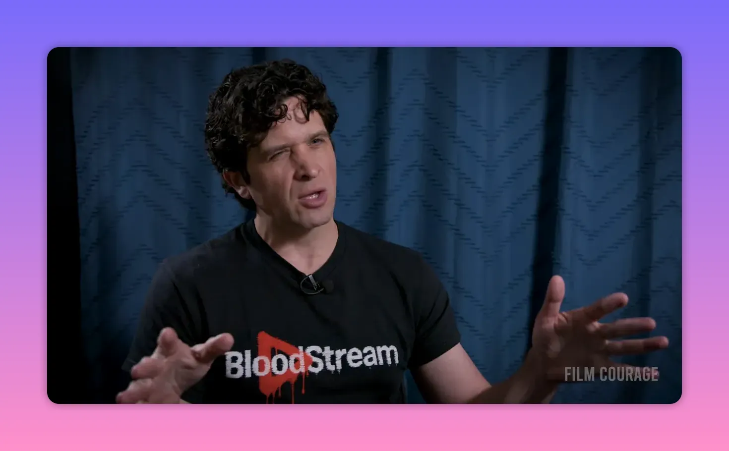 Interviewee in a BloodStream T‑shirt gesturing with both hands while discussing platform scale and partnerships, blue curtain backdrop.