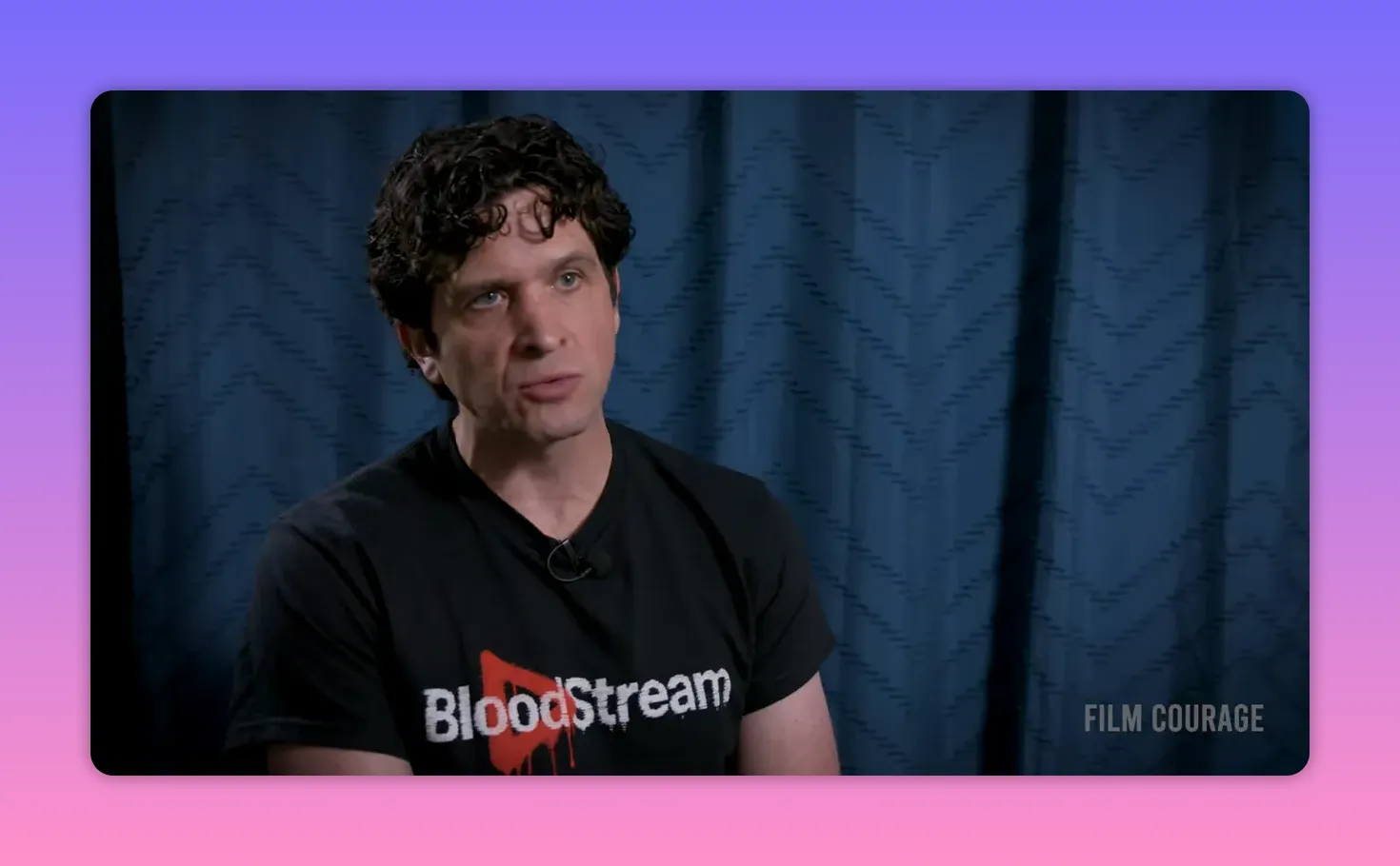 Clear mid-shot of an interviewee wearing a BloodStream T-shirt against a blue curtain, well-lit and composed to show the logo and context.