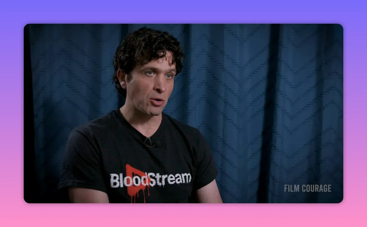 Interview mid-shot of a speaker wearing a BloodStream T‑shirt, engaged and speaking with a blue curtain backdrop; clear, high quality frame.