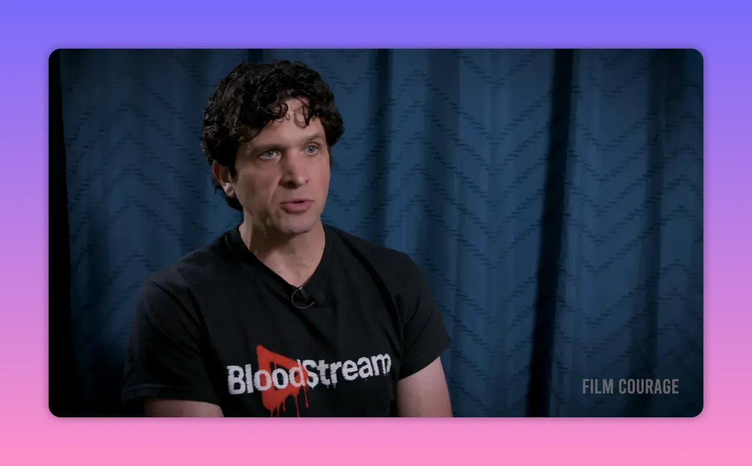 Interviewee wearing a BloodStream t-shirt, engaged expression, clear view of logo against blue curtain.