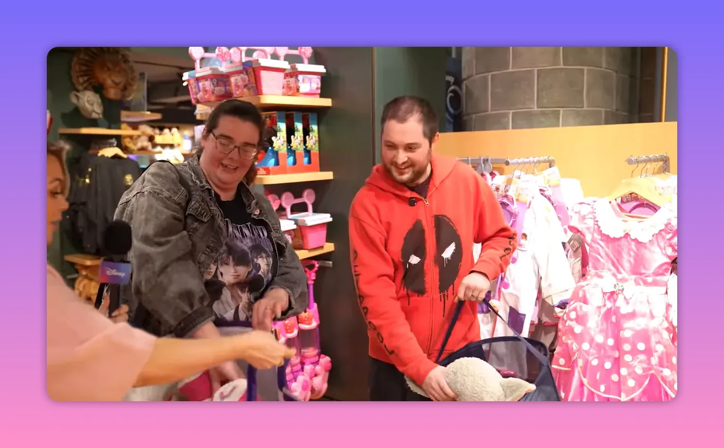Presenter offering a Disney bag while two shoppers prepare to load toys beside costume racks in a Disney Store