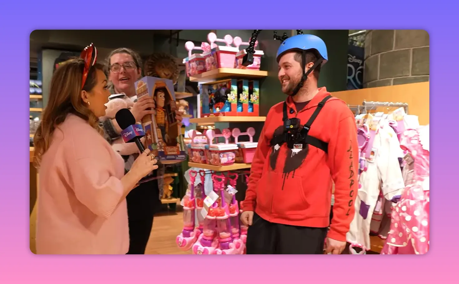 Presenter speaking into a Disney-branded microphone while a helmeted contestant smiles beside shelves of toys