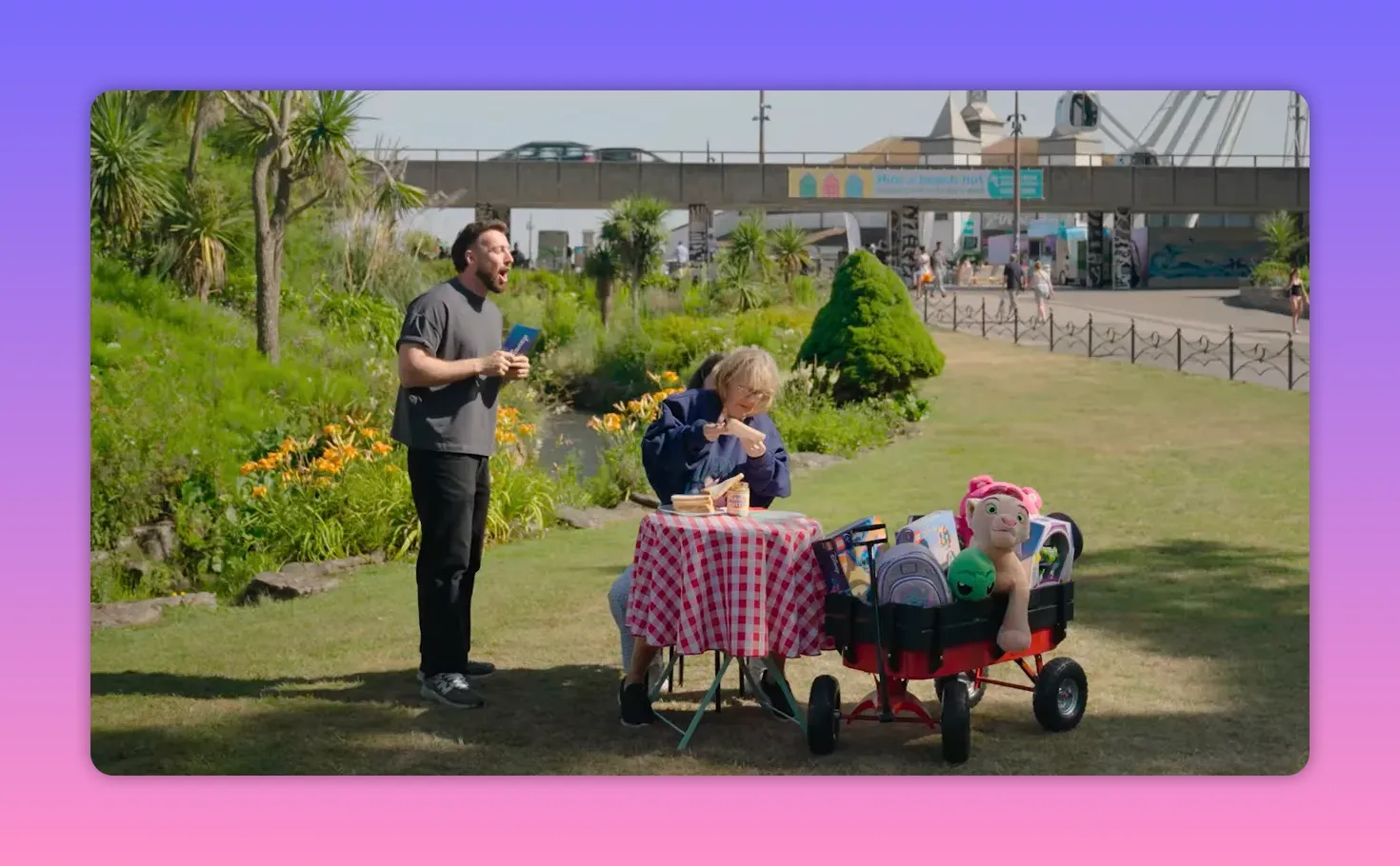 Wide view of host standing next to a contestant at a picnic table with a prize-filled wagon beside them in an outdoor park.