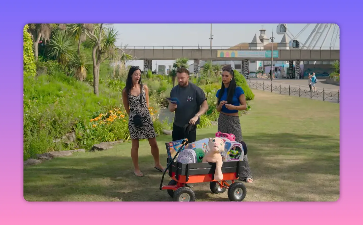 Three contestants stand behind a wagon full of prizes on a grassy lawn; one contestant reacts with surprise while the host reads a card.