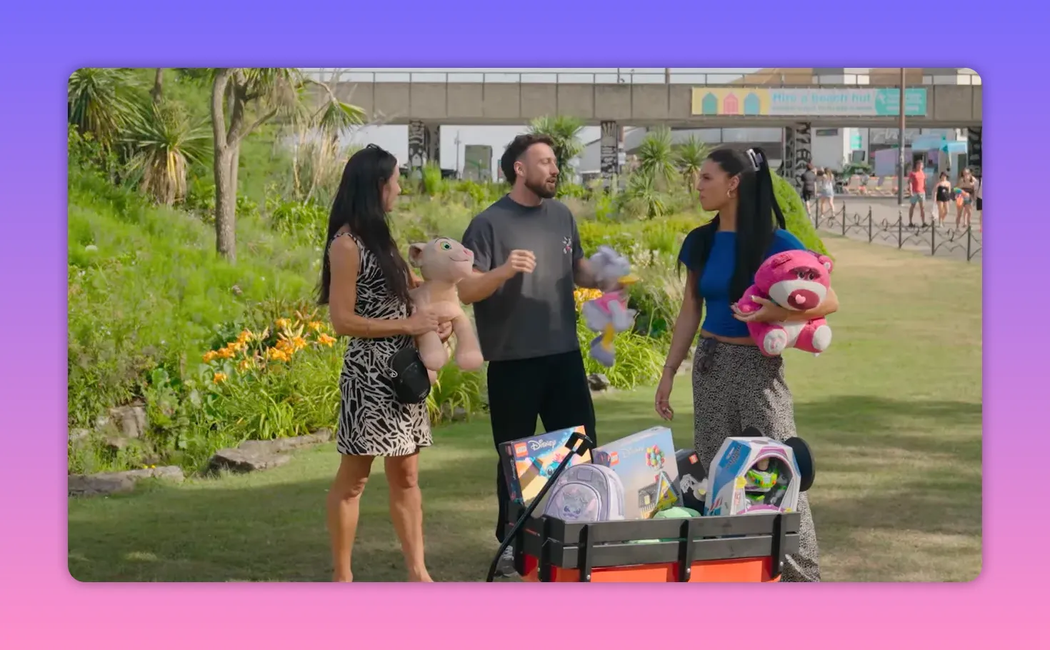 Wide outdoor shot showing a host and two contestants standing behind a small wagon filled with toys and prizes on a grassy area.