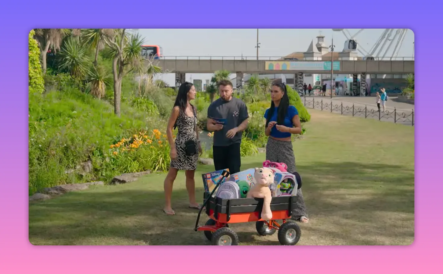 Wide outdoor shot of the host and two contestants standing behind a small red wagon filled with boxed toys and plush prizes on a grassy lawn.