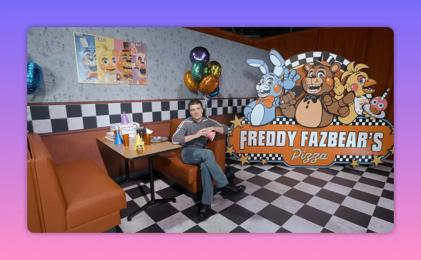 Wide, well-lit shot of the Freddy Fazbear's Pizza set with a person sitting at a booth, colorful balloons, party hats, and a large Freddy Fazbear cardboard sign.