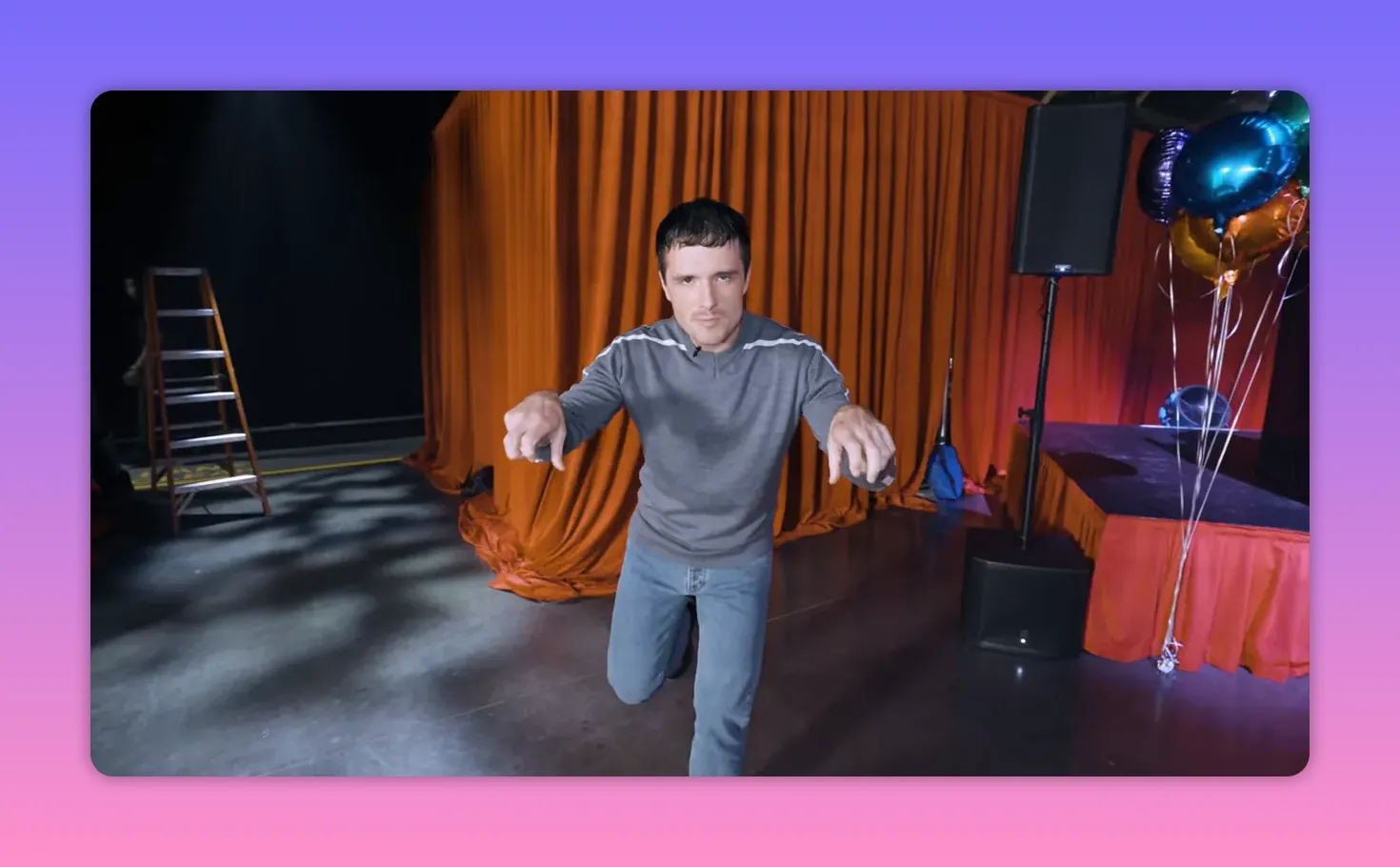 Person leaning forward and pointing toward the camera on a stage with orange curtains, balloons and a speaker visible to the side.