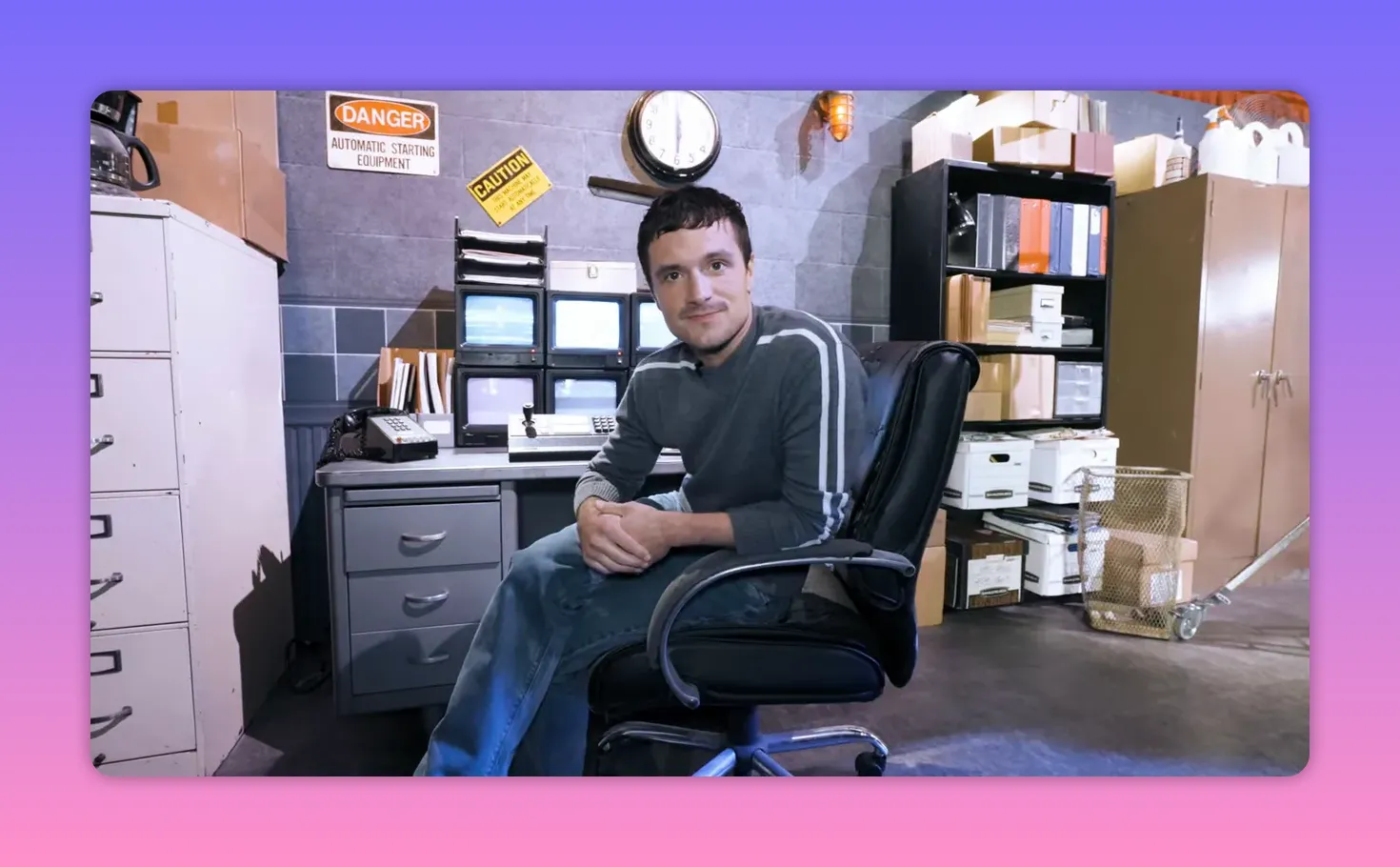 Actor seated at a security office desk on the Fazbear set looking toward the camera, with monitors, filing cabinets and prop boxes behind.