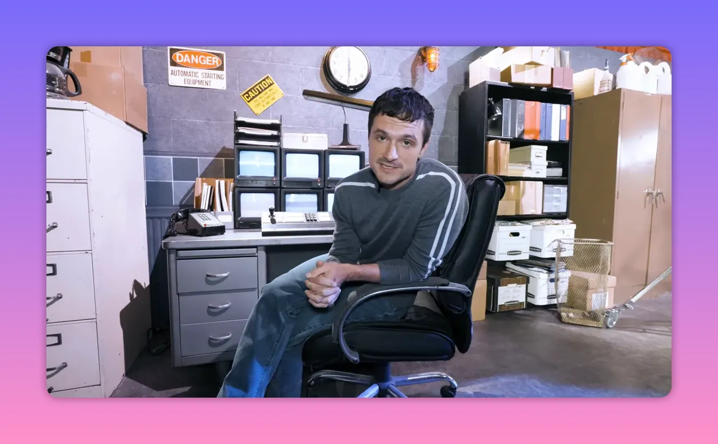 Actor centered in a security-office set sitting in a swivel chair with monitors, filing boxes and a wall clock visible