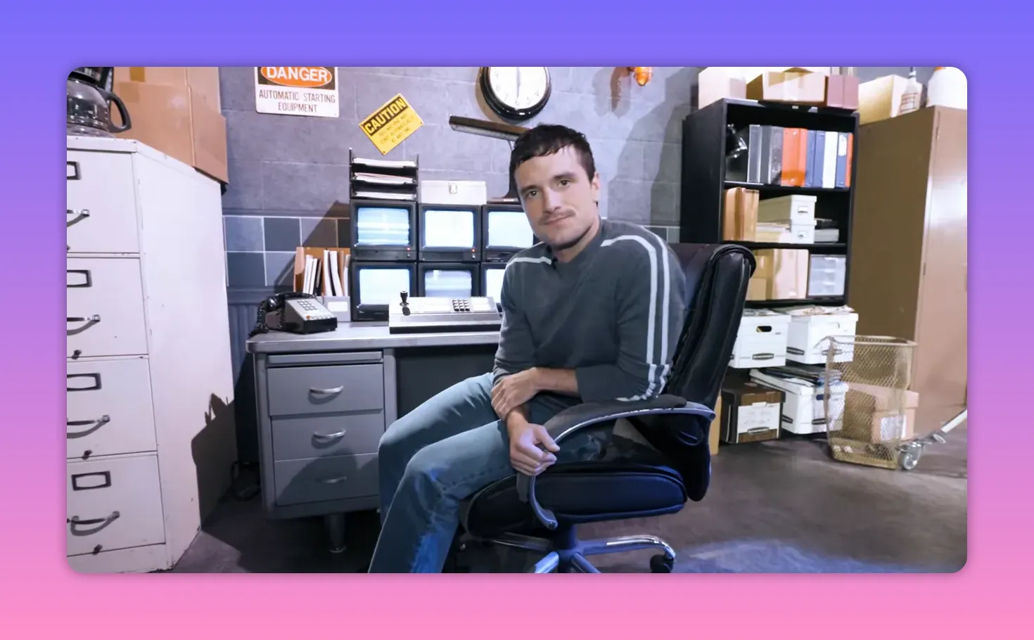 Person seated at a security desk with a bank of small monitors, a wall clock, filing cabinets and storage boxes on the Fazbear set.