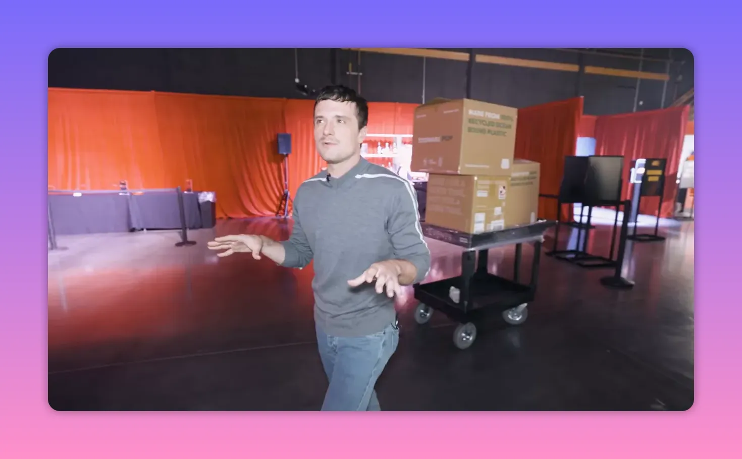 Person walking through an event space with hands extended in mid-gesture and a cart with stacked cardboard boxes behind them; red curtains in the background.