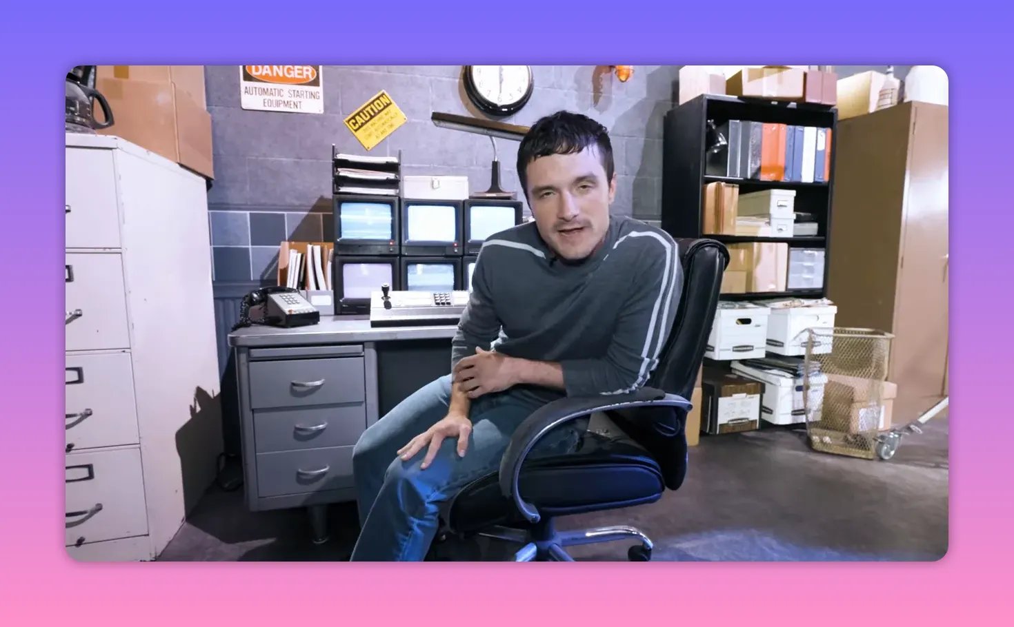 Person seated in a security office set with a bank of monitors, a clock on the wall, and props visible around the desk.
