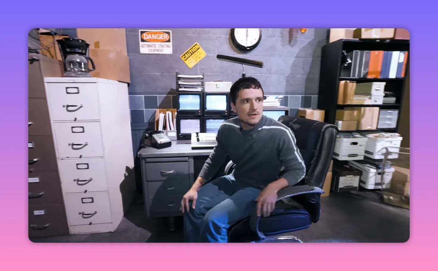 Actor seated in a security office chair surrounded by monitors, filing cabinets, boxes and a wall clock on the Fazbear set.