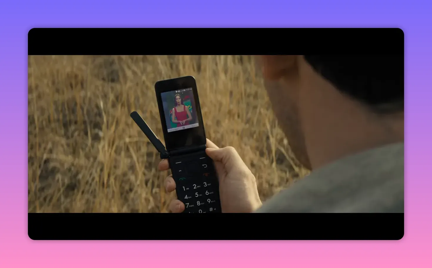 Person holding a flip phone in a dry field viewing a live news weather broadcast on-screen