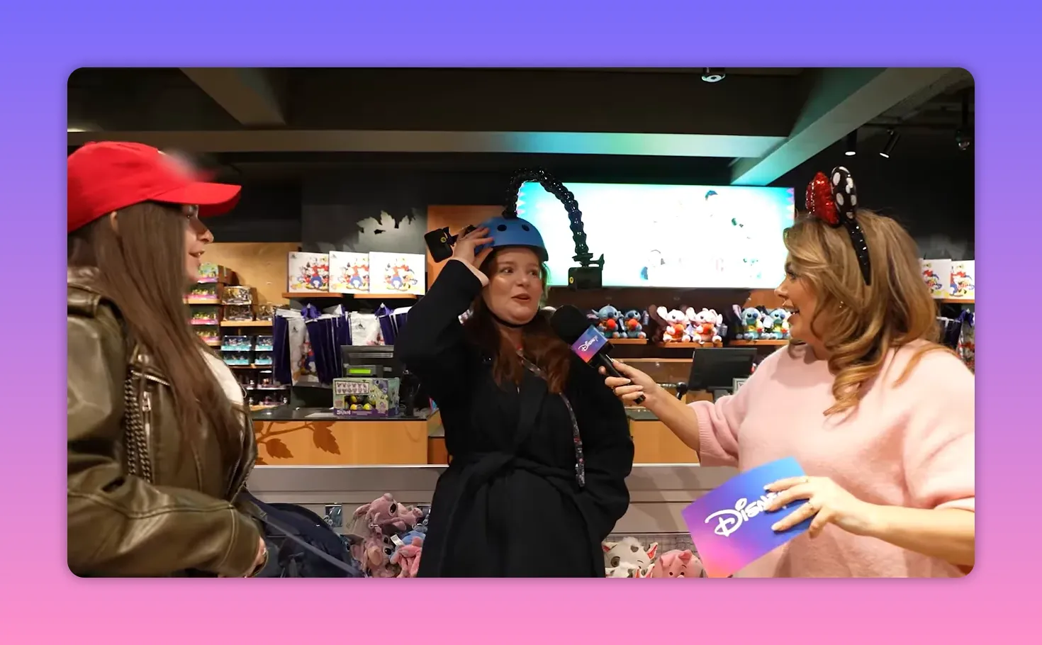 Disney Store host holds a microphone to a contestant wearing a blue helmet with plush displays and a visible prize area behind them.
