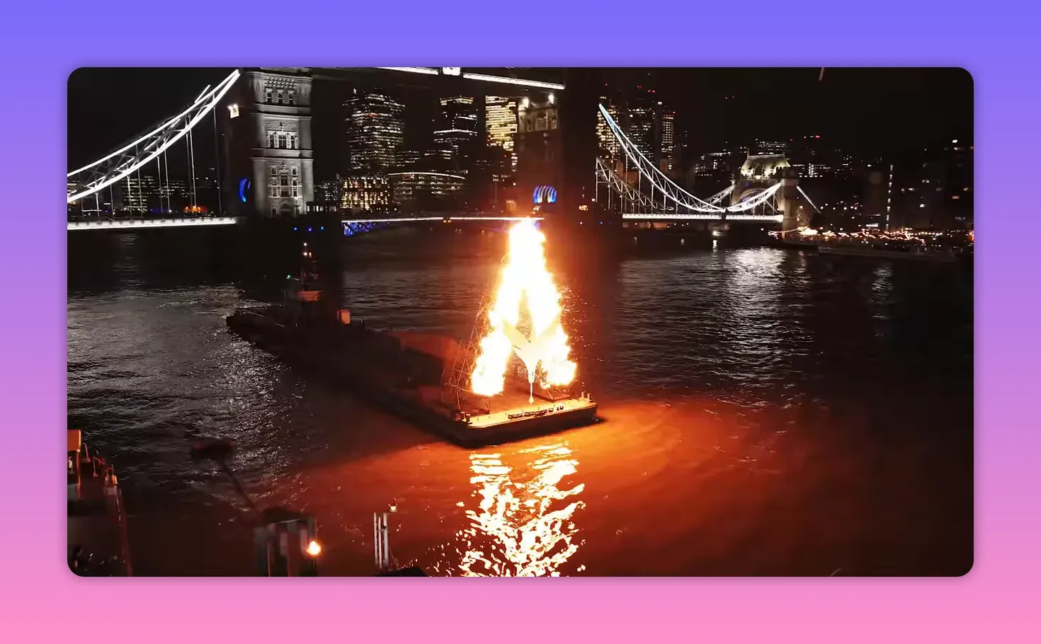 Nighttime view of a large burning 'A' installation on a barge on the River Thames with an illuminated suspension bridge and city skyline behind.