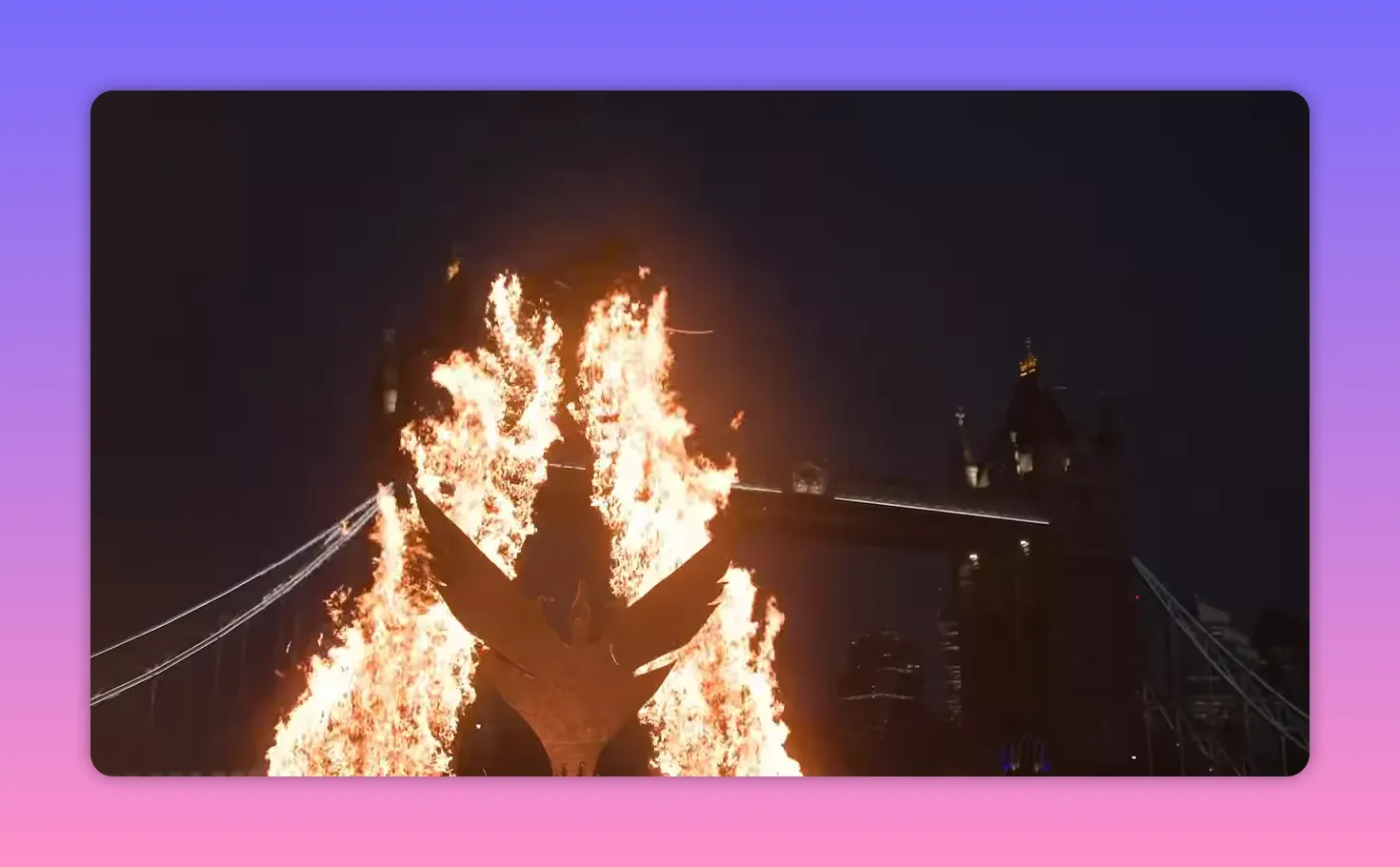 Large flames rising behind a sculptural installation with Tower Bridge faintly visible in the background at night.
