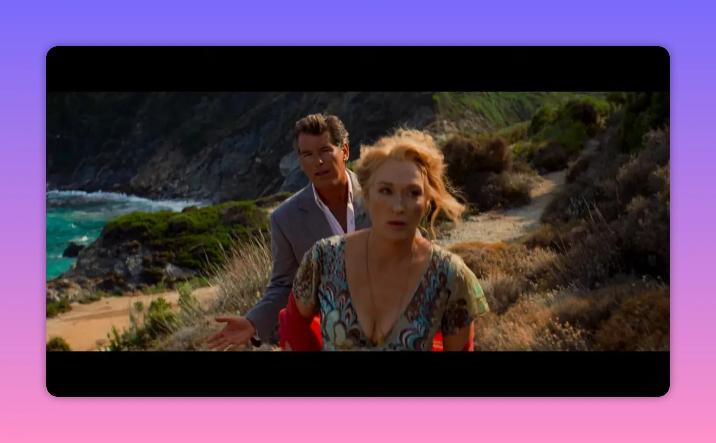 Woman moving forward past a man on a sunlit coastal path with turquoise sea and cliffs in the background.