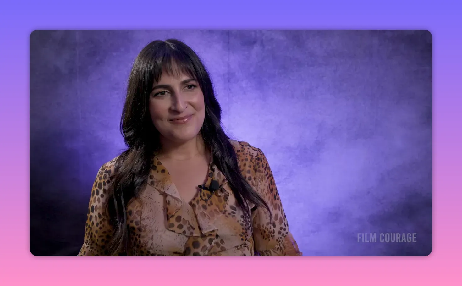 Interview subject looking slightly off-camera with a gentle smile against a purple textured backdrop, wearing a patterned blouse and lapel microphone.