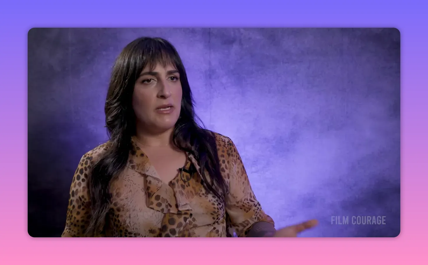 Interview subject gesturing with a hand while speaking against a purple textured backdrop, clear mid-shot with good negative space to the right.