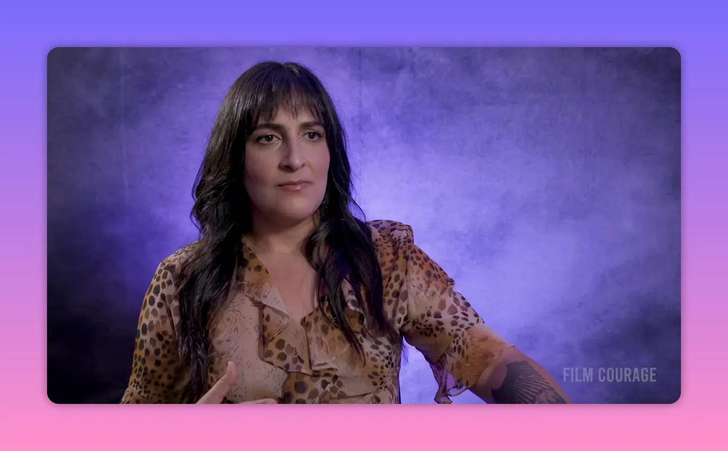 Interview subject seated against a purple textured backdrop with a composed expression and a small hand gesture, clear and well-lit.