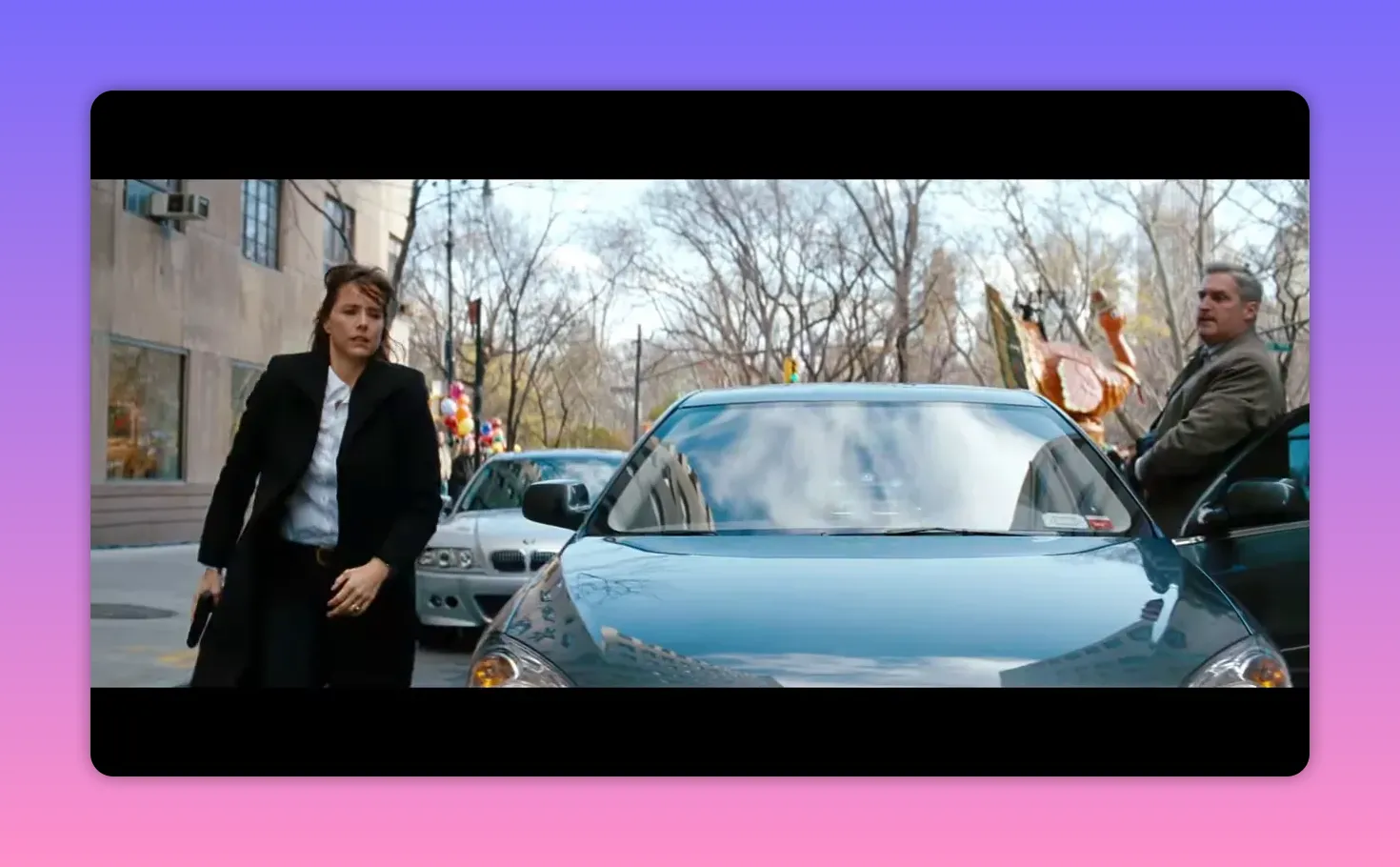 Wide view of a parked car on a city street with parade balloons in the background and a person walking beside it