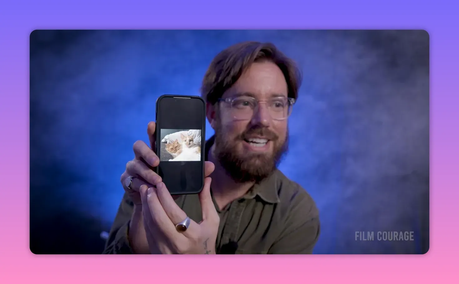Filmmaker holding a smartphone upright showing a clear photo of two cats on the screen, smiling in the background.