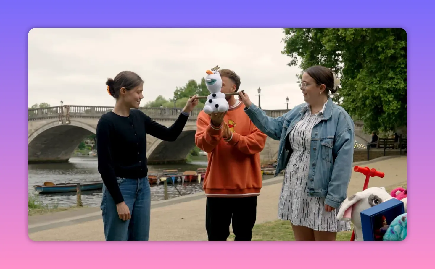 Balanced wide-medium shot of two teammates and the host holding a plush Olaf by its arms beside a river and prize cart, showing the synchronization moment.