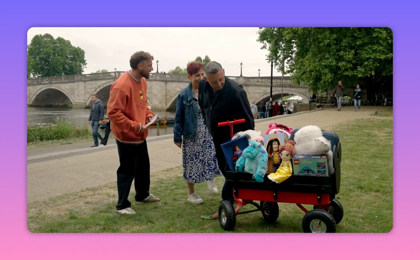 Prize-filled cart with Disney toys and a host speaking to participants by a riverside bridge.