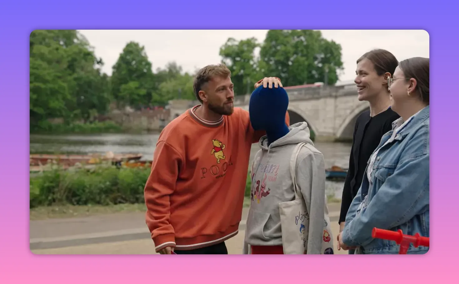 Host leaning in toward a mannequin while two smiling contestants listen beside a riverside path, showing a friendly, engaged hosting moment.