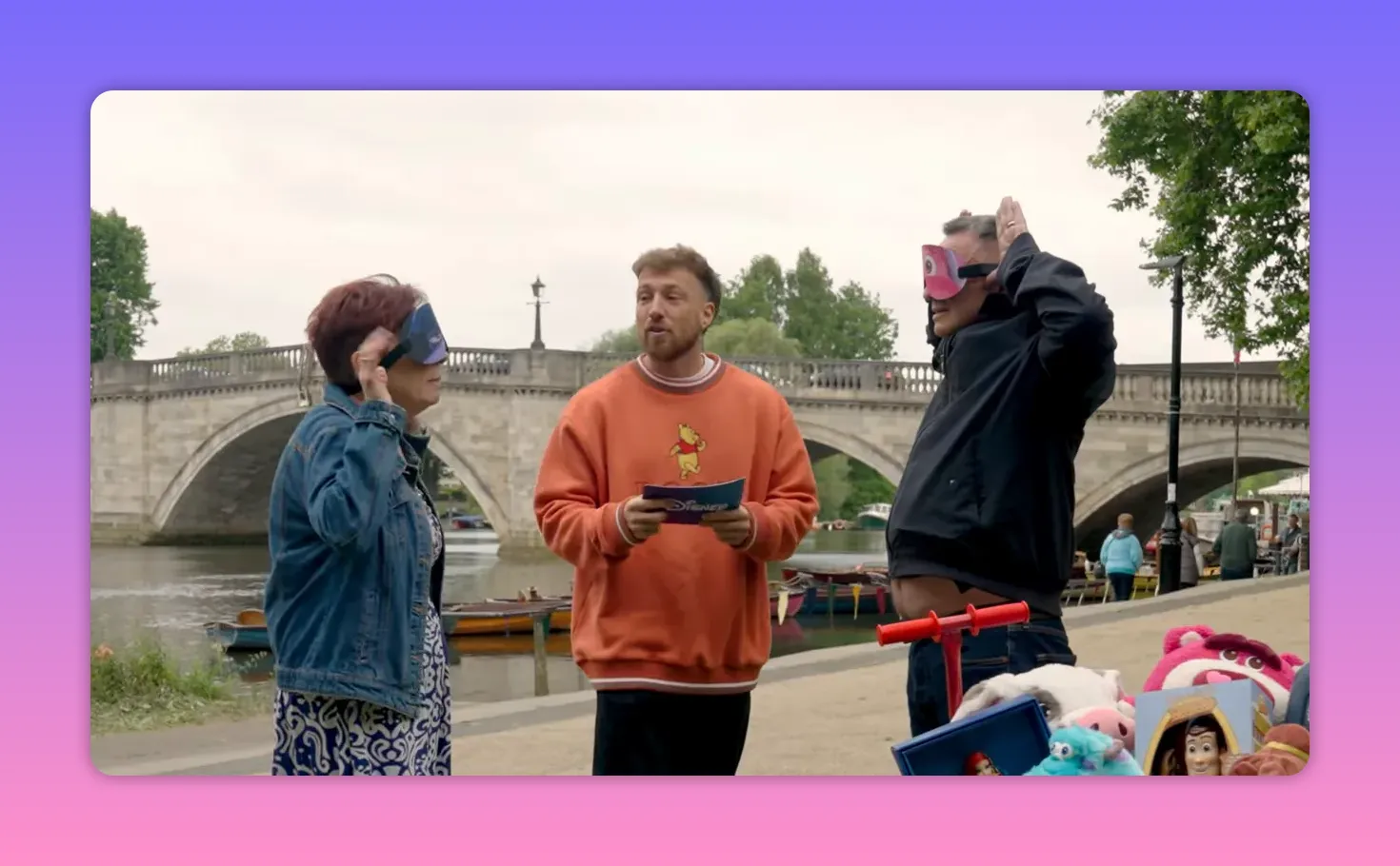 Host standing between two contestants who are putting on blindfolds, with a riverside bridge and prizes visible in the foreground.