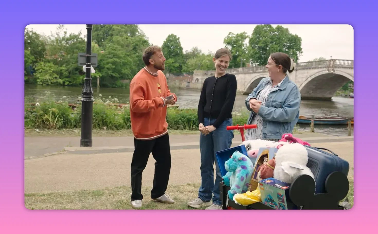 Wide shot of the host speaking to two contestants with a prize-filled cart and stone bridge visible behind them at the riverbank.