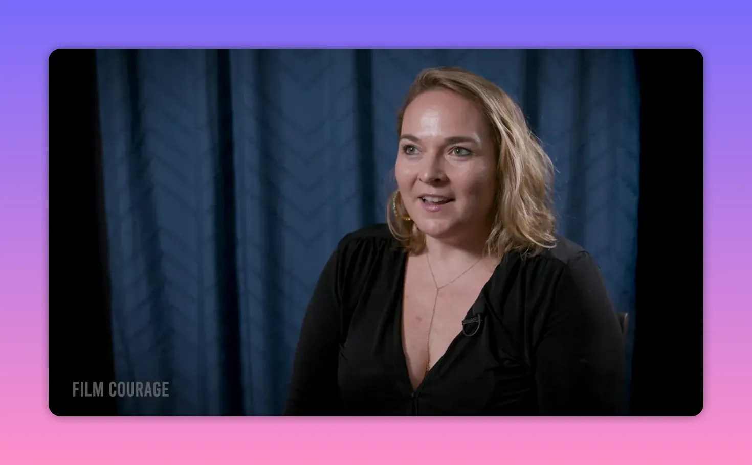 Well-lit mid-shot of a filmmaker speaking thoughtfully, making direct eye contact, seated against a blue curtain.
