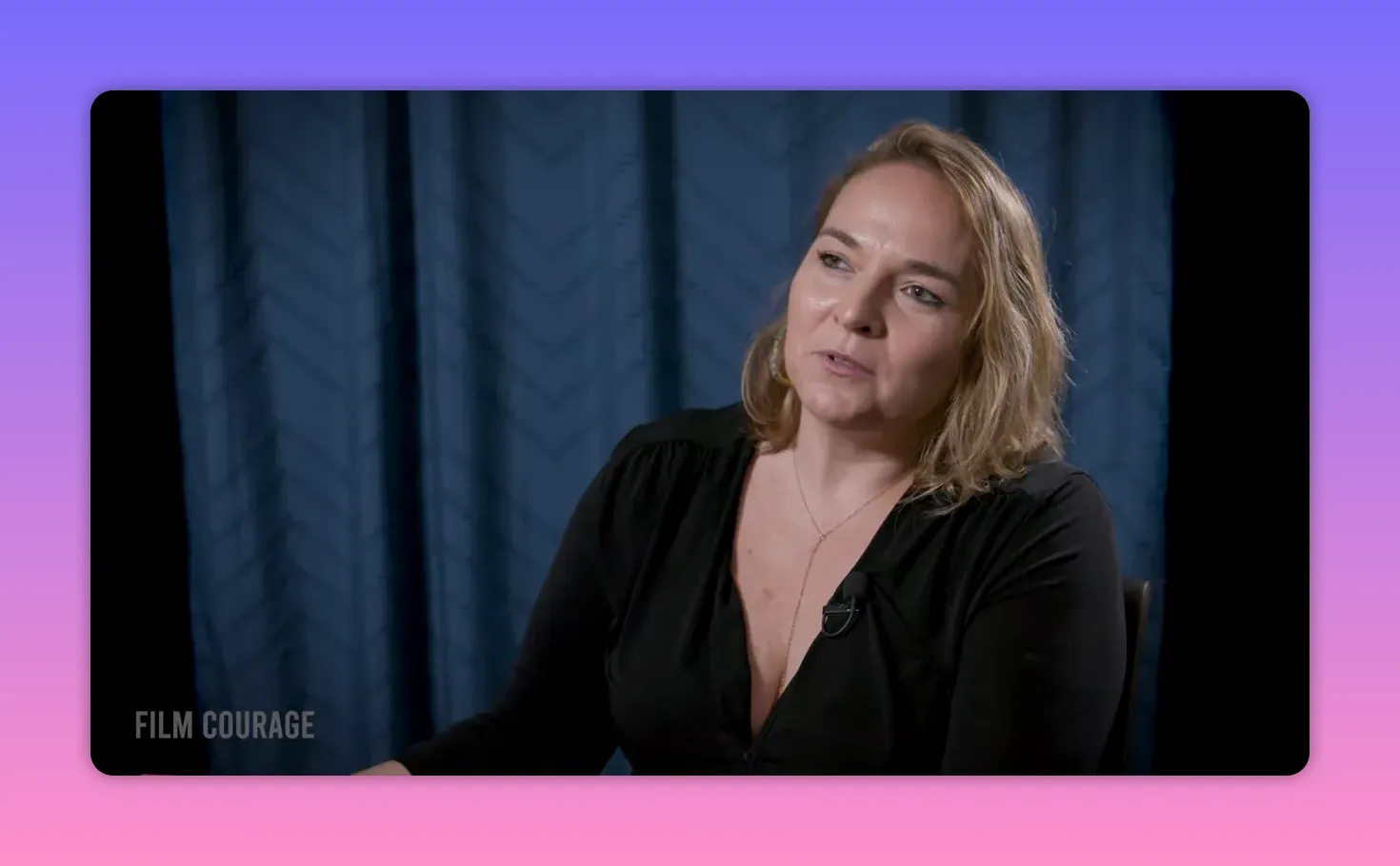 Medium-shot interview of the speaker with a slight smile, seated against a blue curtain and speaking thoughtfully.