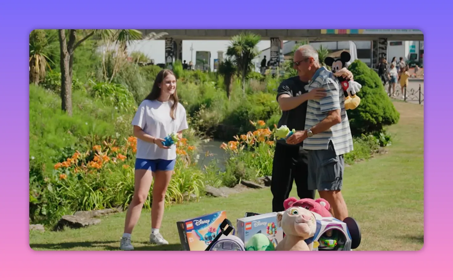Host placing a Mickey plush on the grandfather's shoulder while the granddaughter smiles, toys and boxed prizes in front