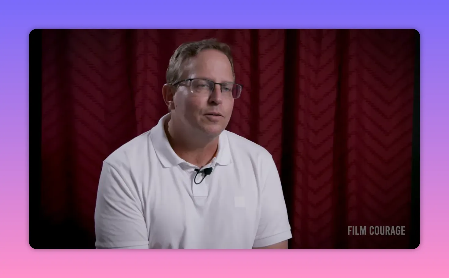 Well-composed interview portrait of a man in a white polo and glasses against a maroon curtain, reflective expression and clear lighting, Film Courage watermark
