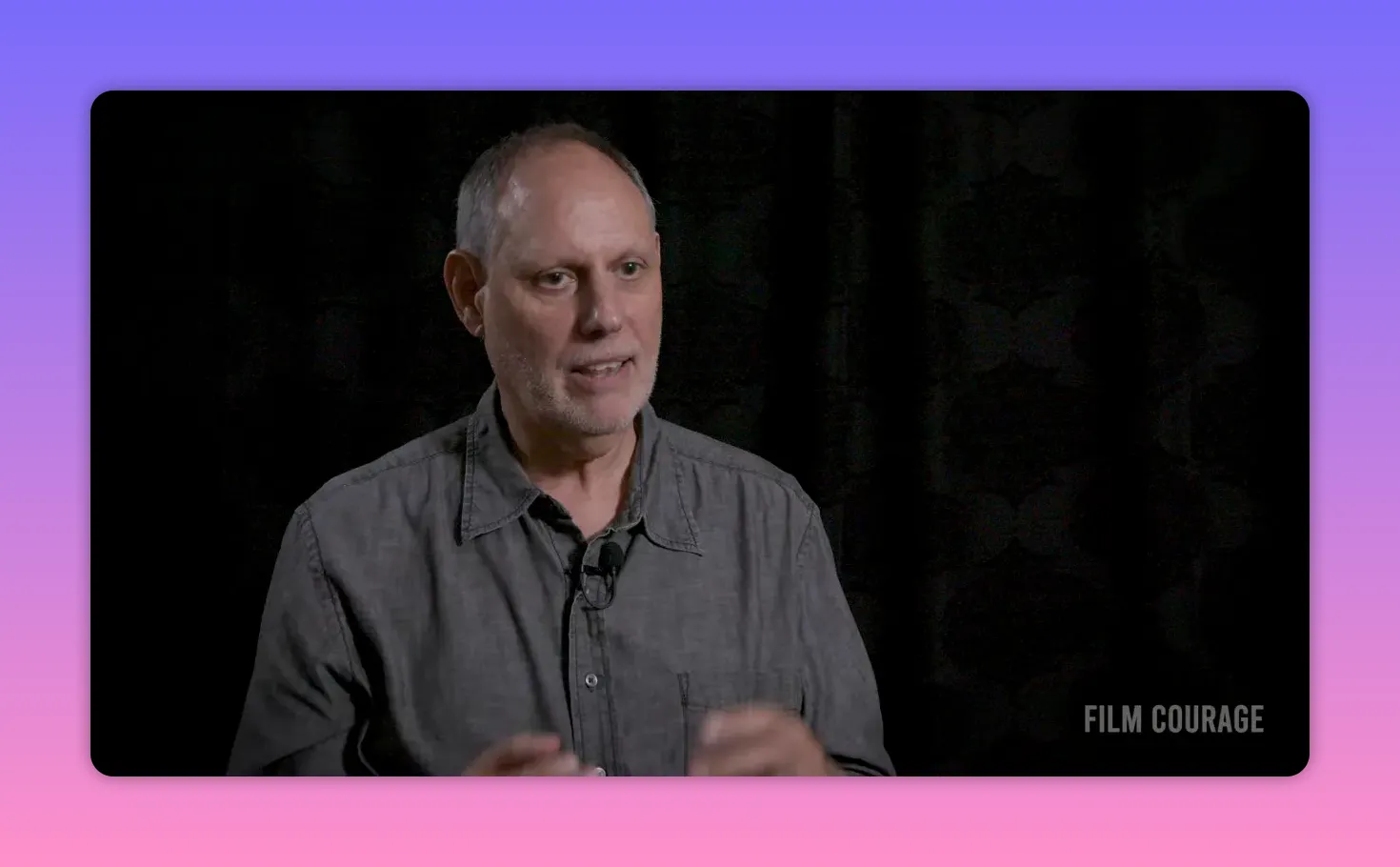 on-camera interviewee in gray shirt against dark patterned backdrop, medium shot with hands visible