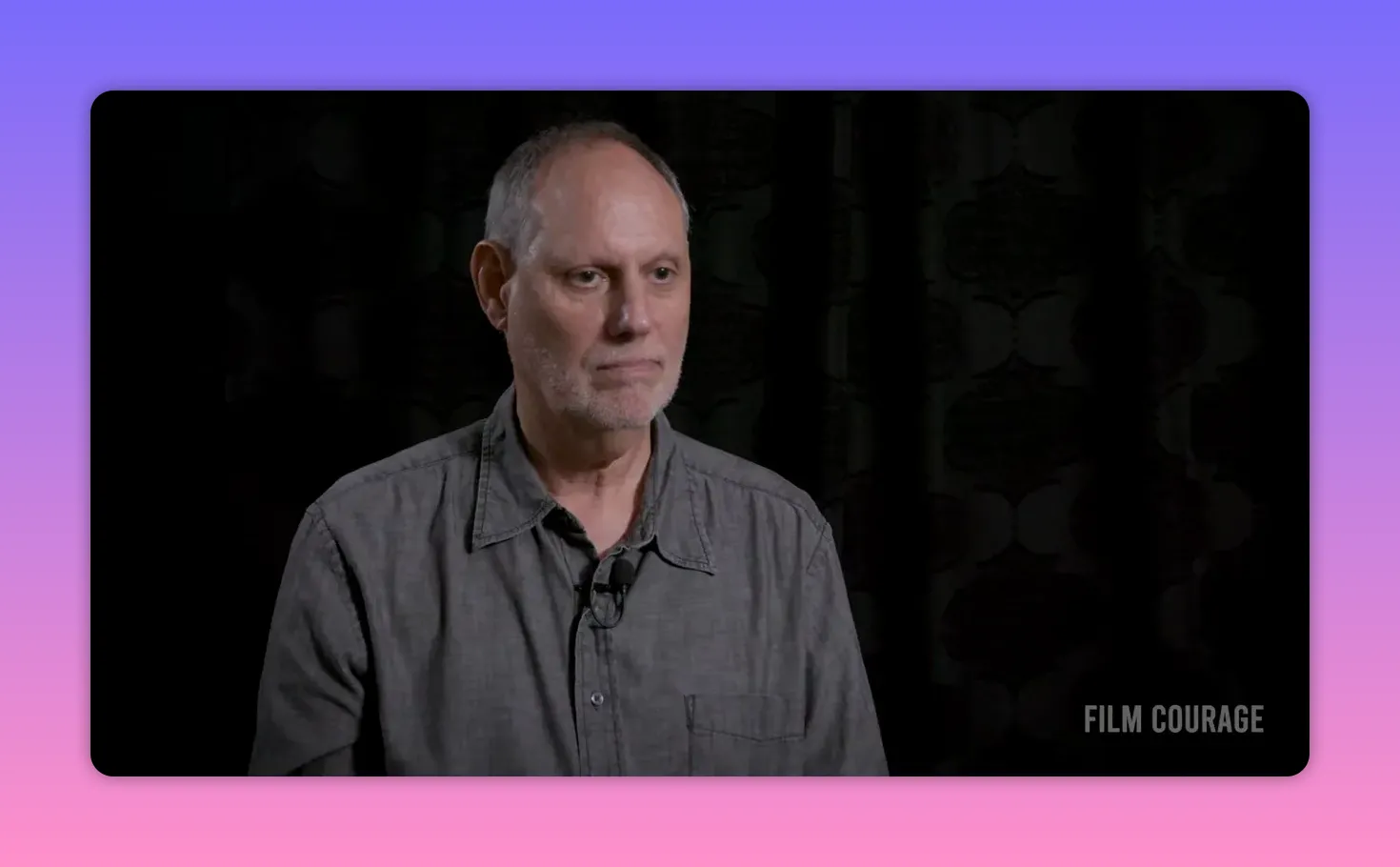 Interviewee in a gray shirt looking calmly forward in a clear medium shot, dark patterned backdrop.