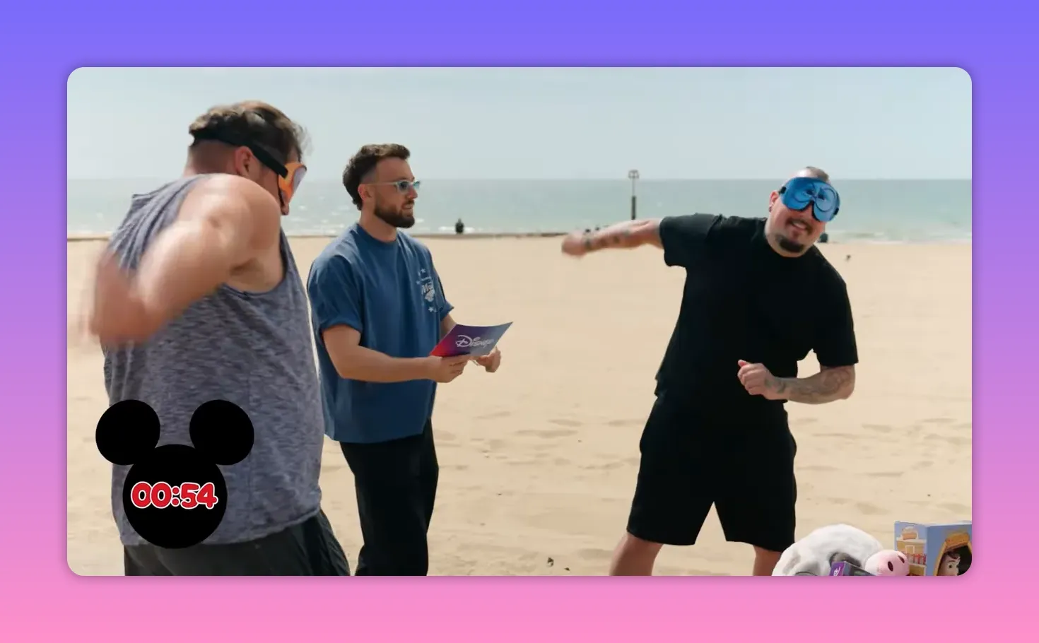 Two blindfolded players performing matching gestures while the host watches on the beach.