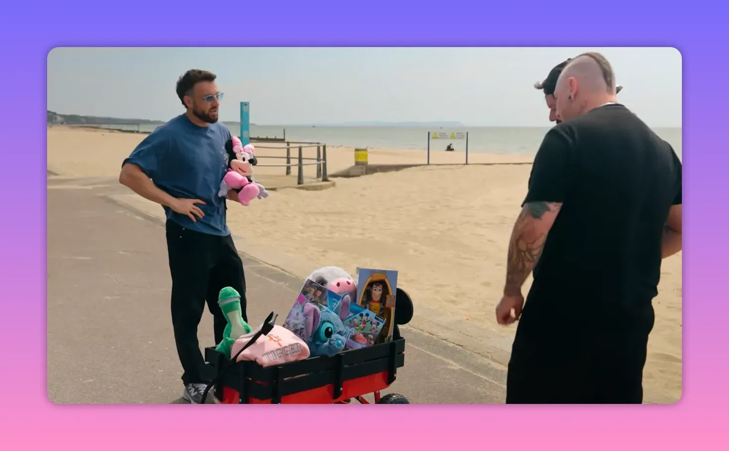 Host holding a Minnie Mouse plush beside a wagon full of Disney toys on the beach promenade with the sea in the background.