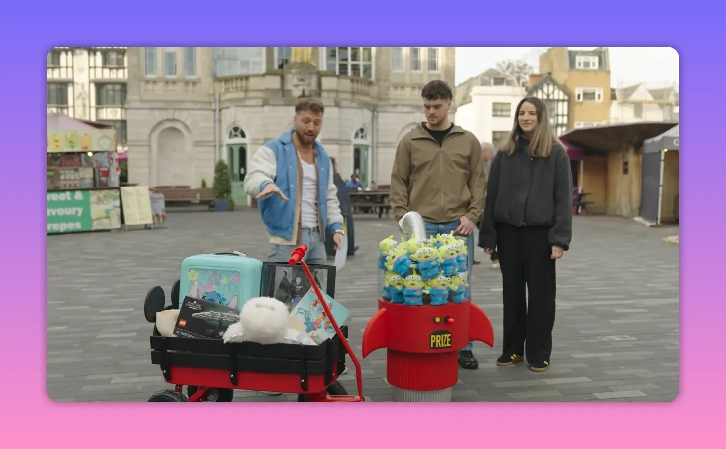 Host and two contestants standing behind a wagon of prizes and a prize dispenser filled with plush toys