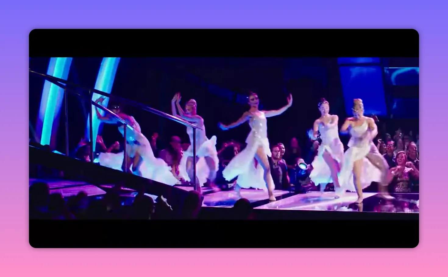 Several dancers in flowing white costumes performing on a brightly lit stage with audience members visible at the edge of the runway.