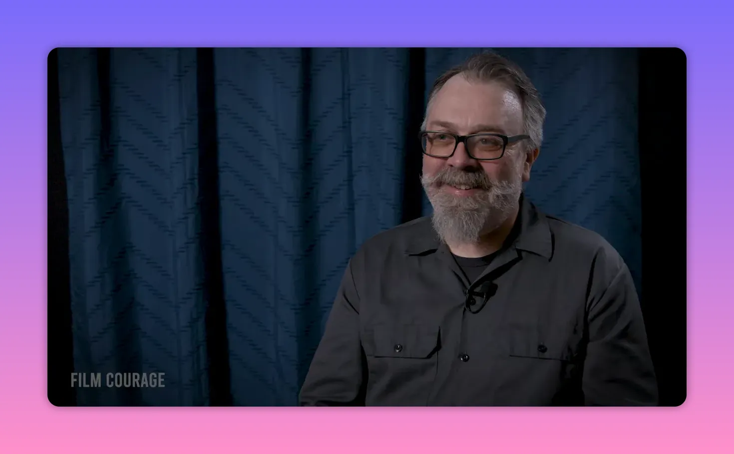 Bearded man smiling during an interview, well-lit, seated in front of a blue curtain.