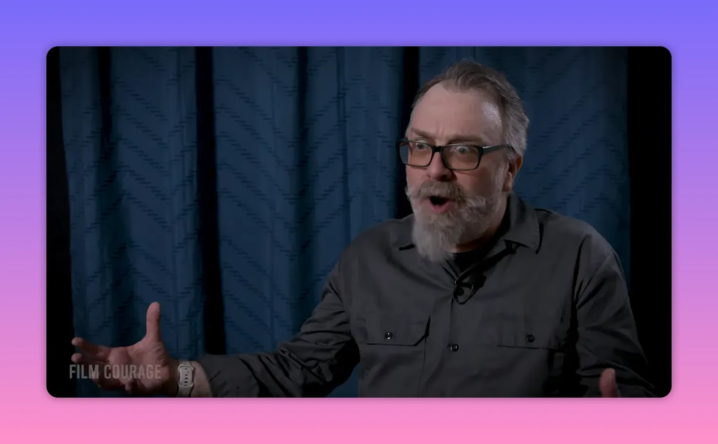Bearded man with glasses making an expressive open-handed gesture while speaking against a blue curtain backdrop
