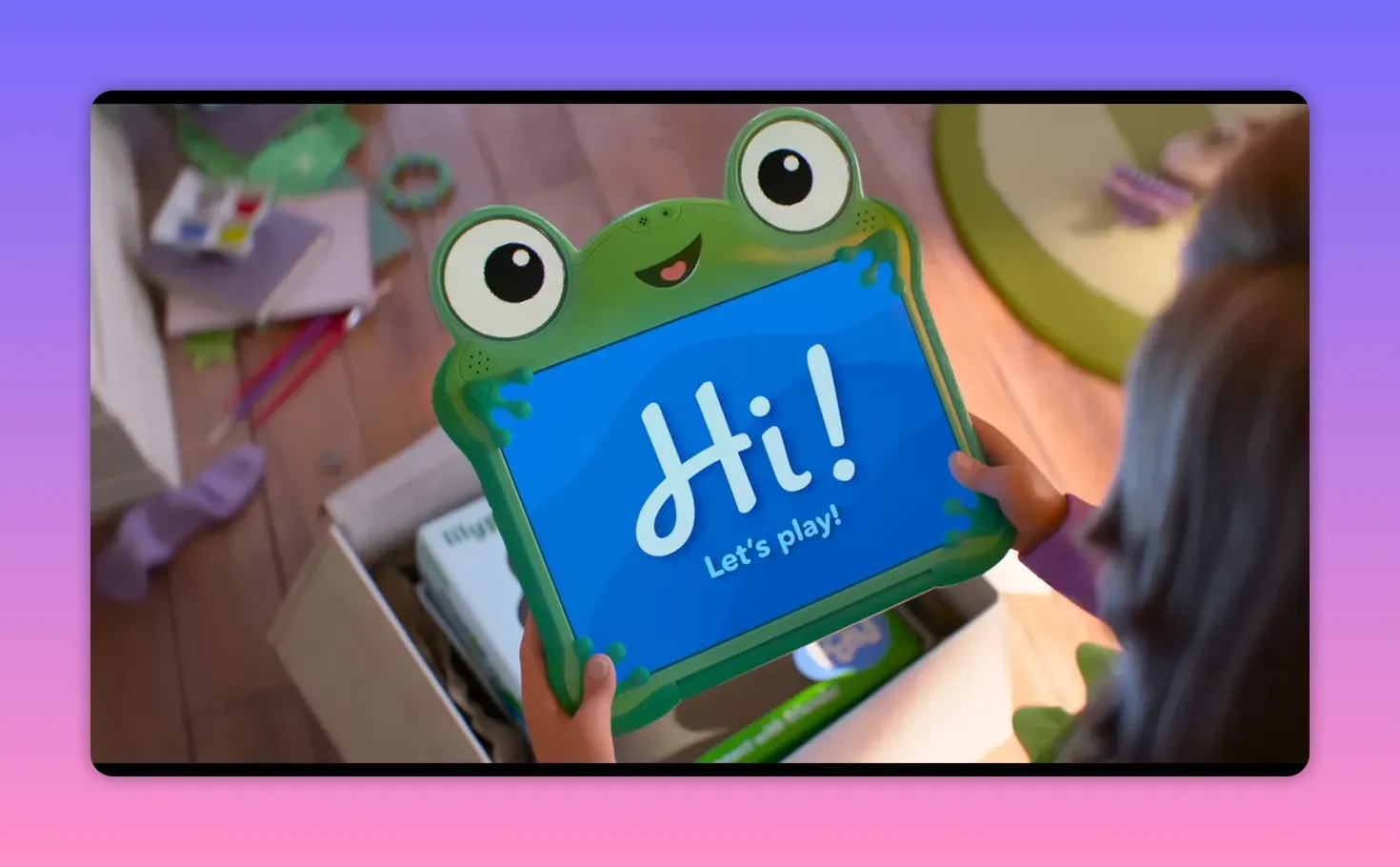 Child holding a frog-shaped tablet that displays the message 'Hi! Let's play!' above an open box.