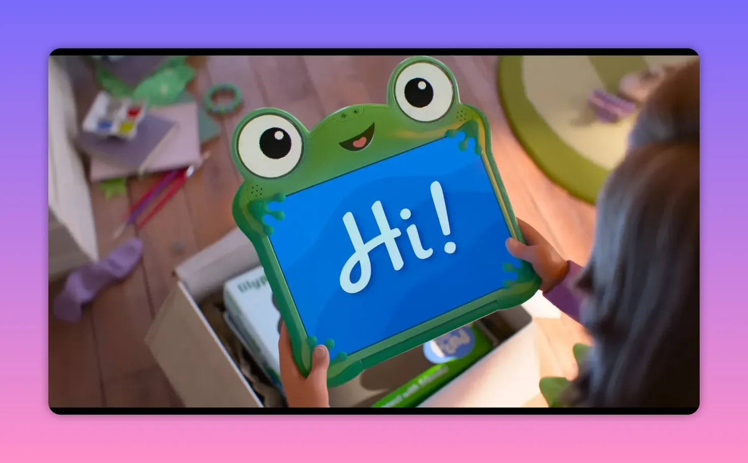 Frog-shaped tablet displaying the word 'Hi!' being held above an open package in a child's room.