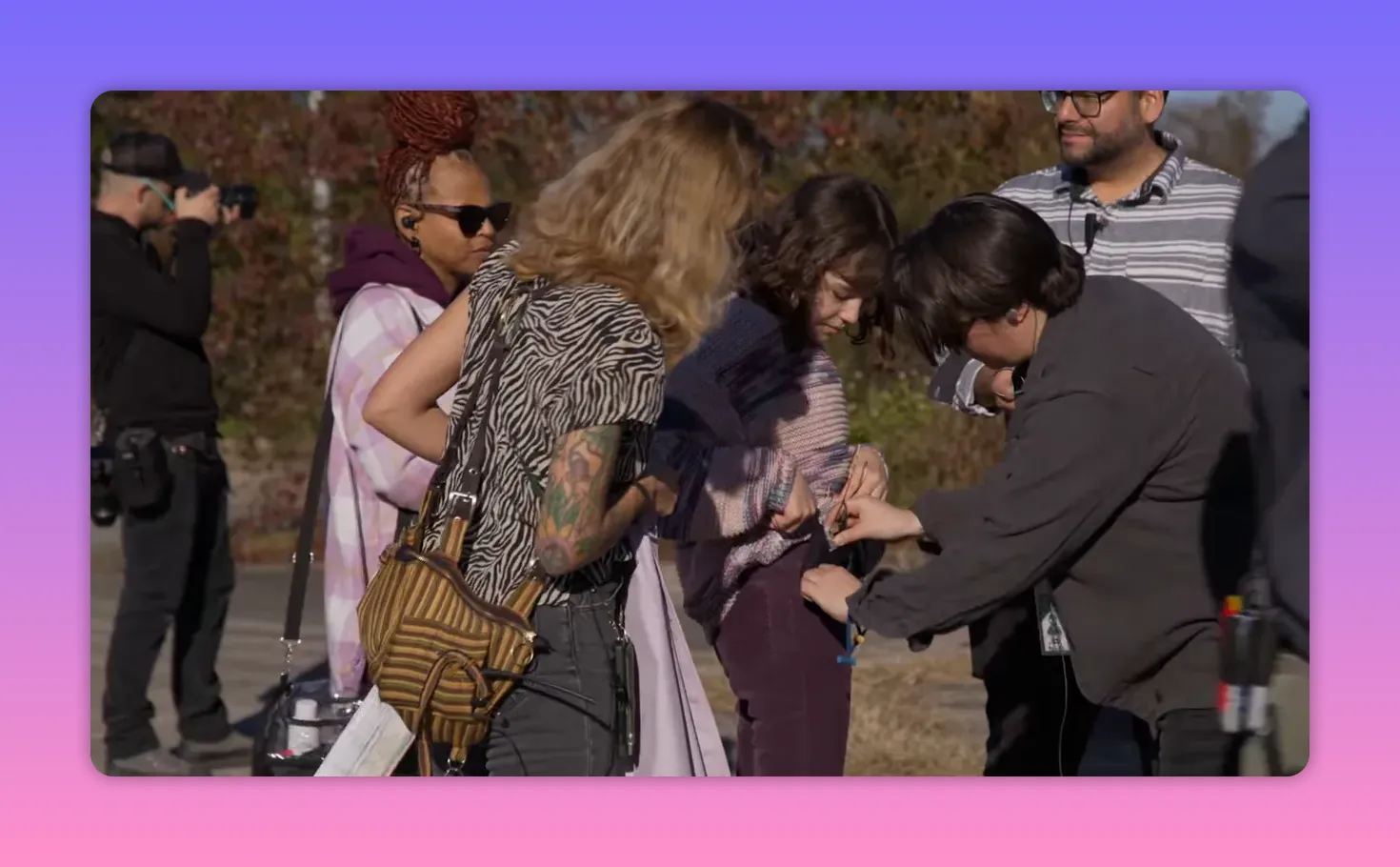 Wardrobe and sound crew adjust an actor's costume and microphone at an outdoor rehearsal, hands clipping and fastening a mic to the actor's waist.