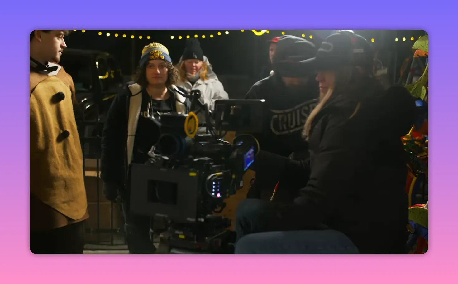 On-set wide shot showing a camera rig, crew and several costumed extras under string lights during a night shoot.