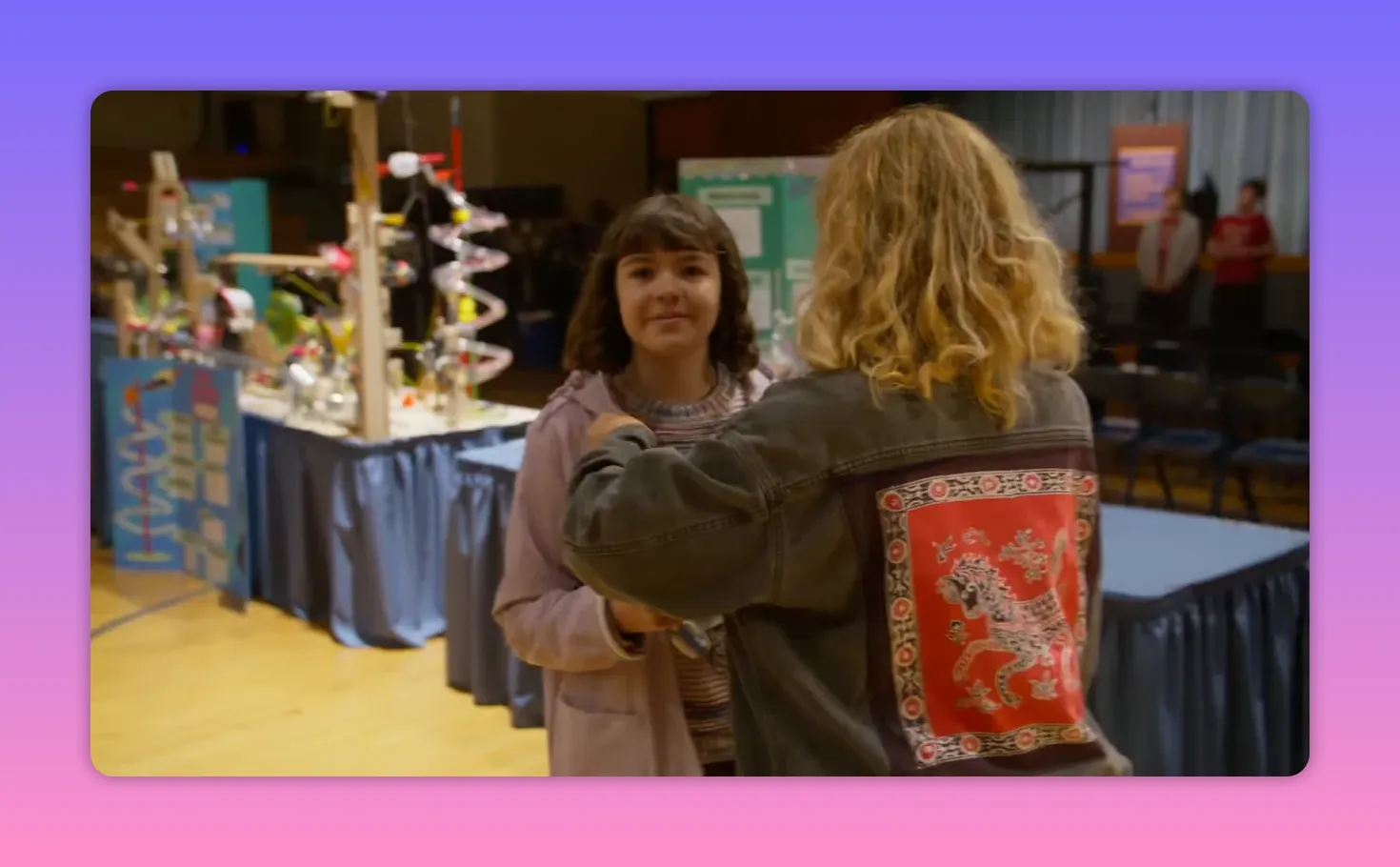 Costumer fitting a microphone or adjusting a jacket on a young actor in a gym-like set.